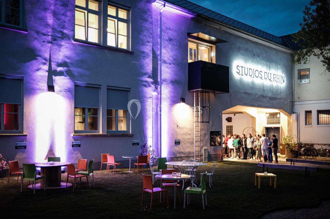 Façade éclairée des Studios du Rhin avec des tables et chaises colorées sur une pelouse, de nuit.