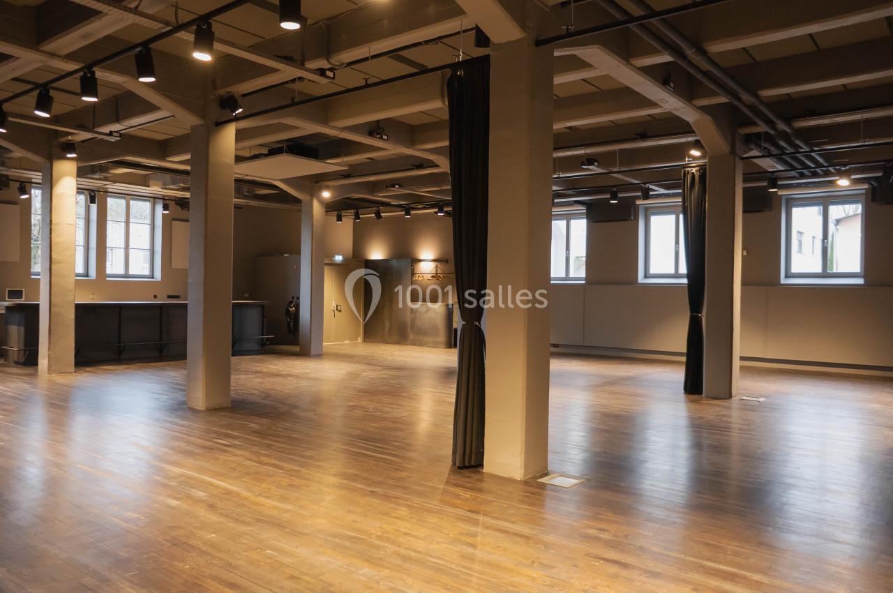 Salle spacieuse avec parquet en bois, colonnes grises, rideaux noirs et éclairage suspendu, baignée de lumière naturelle.