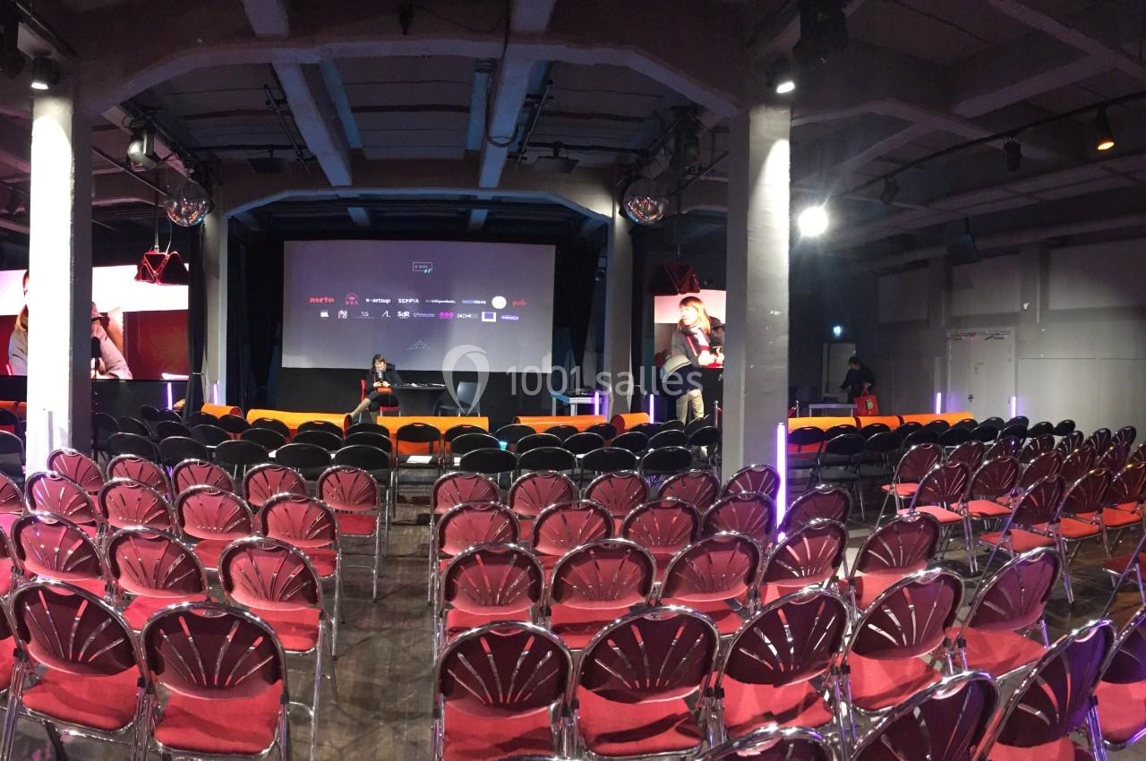 Salle de conférence vide avec rangées de chaises rouges, éclairage de scène et écran de projection en arrière-plan.