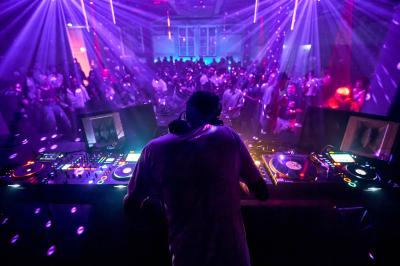 Un DJ mixe sur une scène éclairée par des lumières colorées, face à une foule dansante dans une salle sombre.