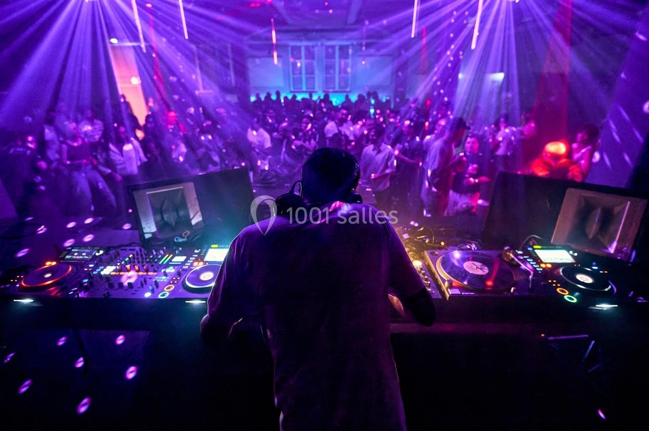 Un DJ mixe sur une scène éclairée par des lumières colorées, face à une foule dansante dans une salle sombre.