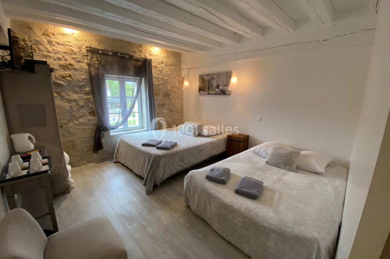 Chambre lumineuse avec deux lits doubles, murs en pierre apparente, fenêtre avec rideaux et mobilier simple.