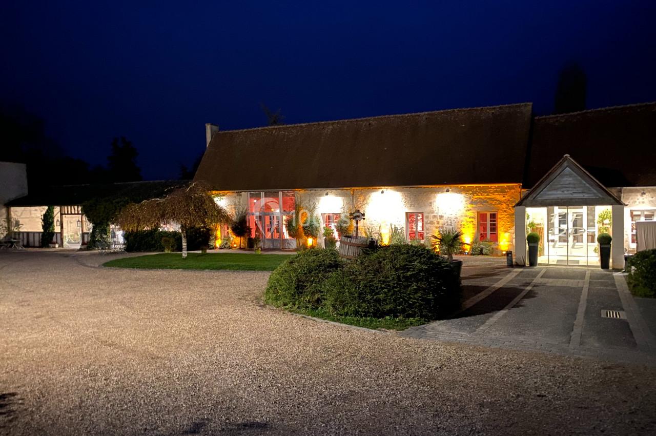 Bâtiment rural éclairé de nuit avec des murs en pierre, des fenêtres rouges et une entrée principale illuminée.