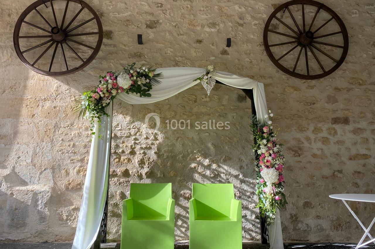 Arche de mariage décorée de fleurs blanches et roses, avec deux chaises vertes devant un mur en pierre.