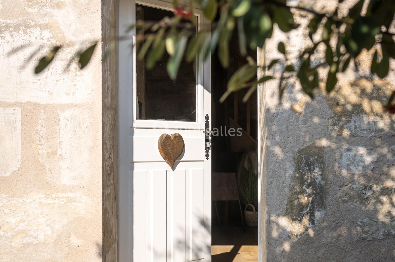 Porte blanche entrouverte avec un cœur en bois, encadrée par un mur en pierre et des branches avec des baies rouges.