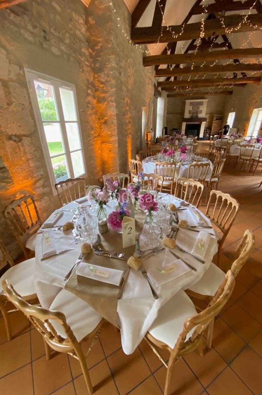Salle de réception décorée avec des tables rondes dressées, ornées de fleurs et éclairées par des guirlandes lumineuses.