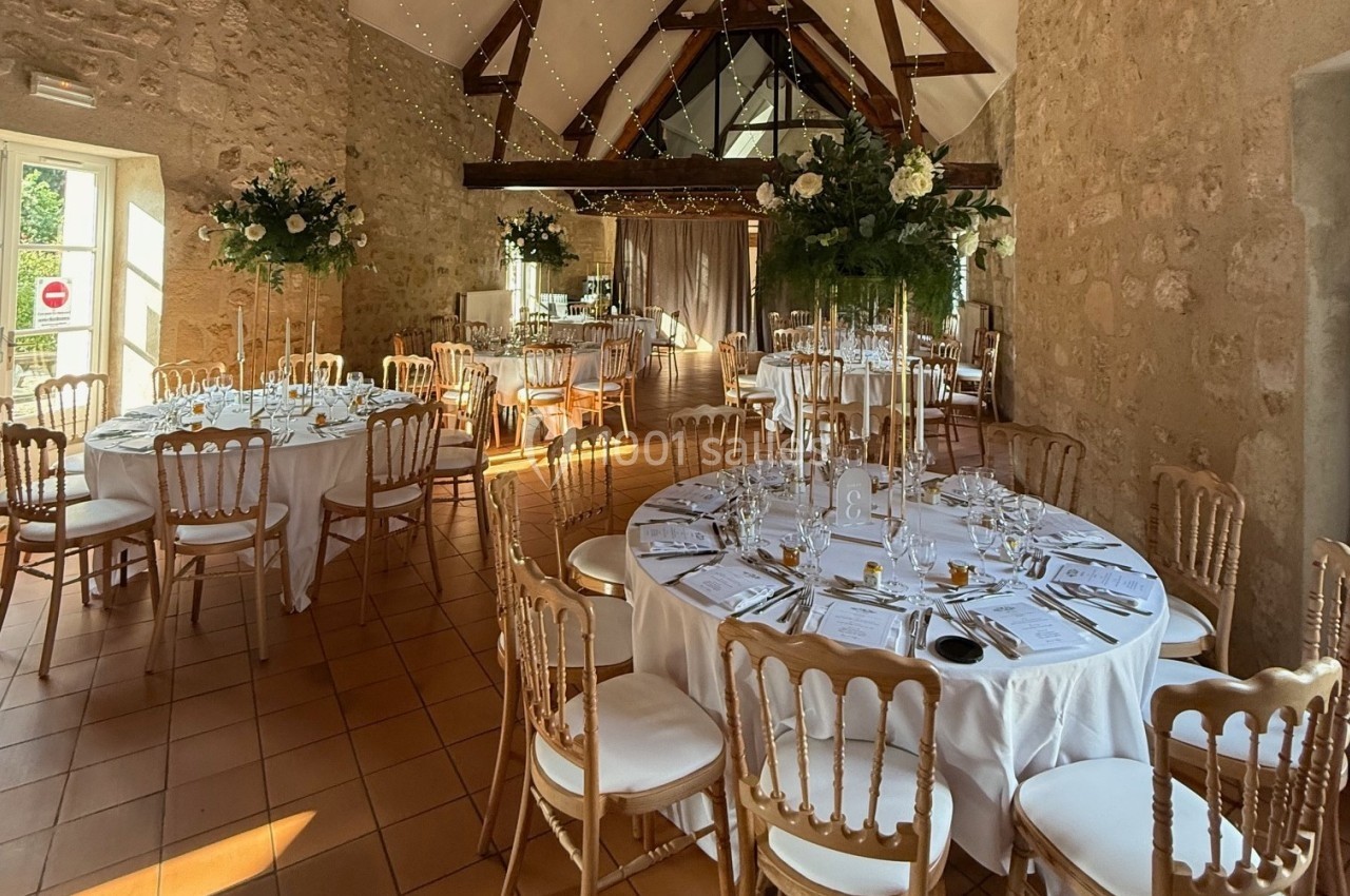 Salle de réception décorée pour un mariage avec tables rondes dressées, chaises dorées et arrangements floraux.