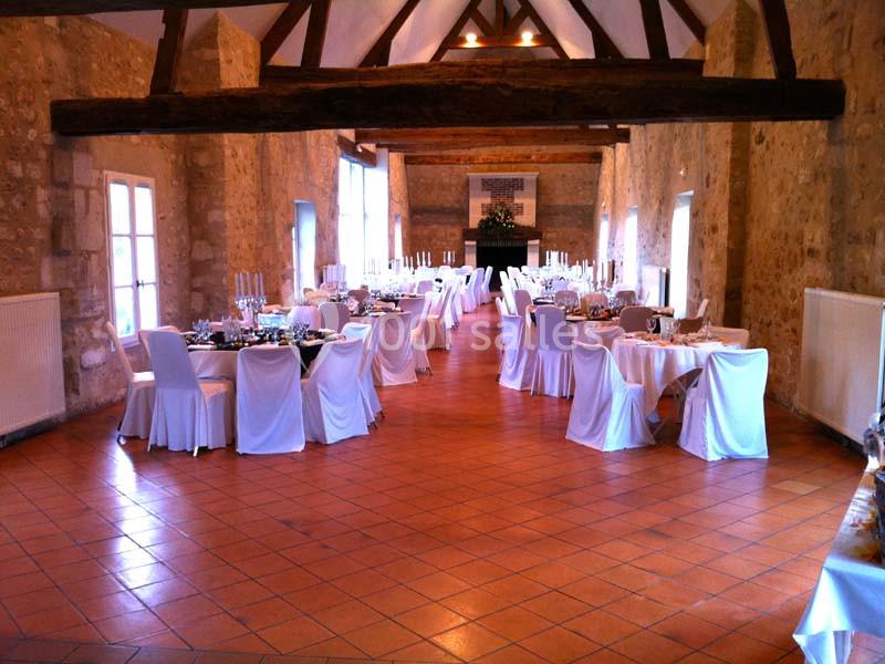 Salle de réception avec des murs en pierre, des tables dressées et des chaises blanches sous une charpente en bois.