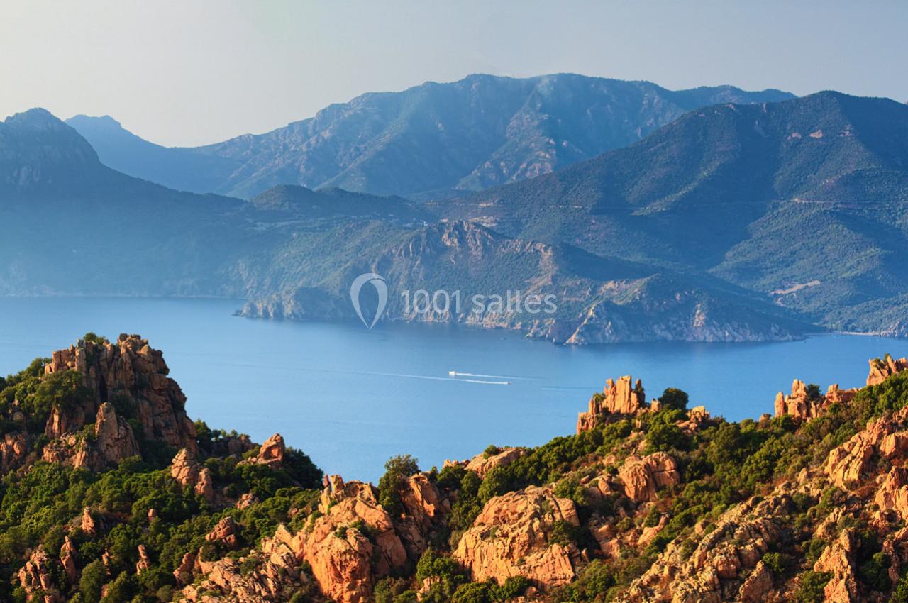 Paysage côtier avec des formations rocheuses rouges au premier plan, une mer bleue et des montagnes verdoyantes à l'arrière…