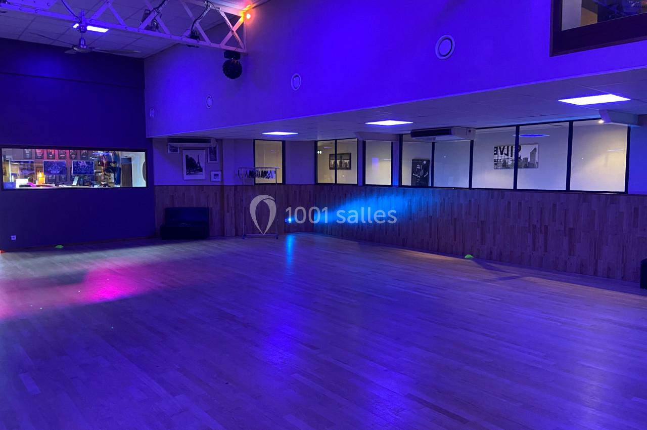 Salle de danse vide avec parquet clair, éclairage tamisé bleu et violet, et murs vitrés donnant sur un espace adjacent.