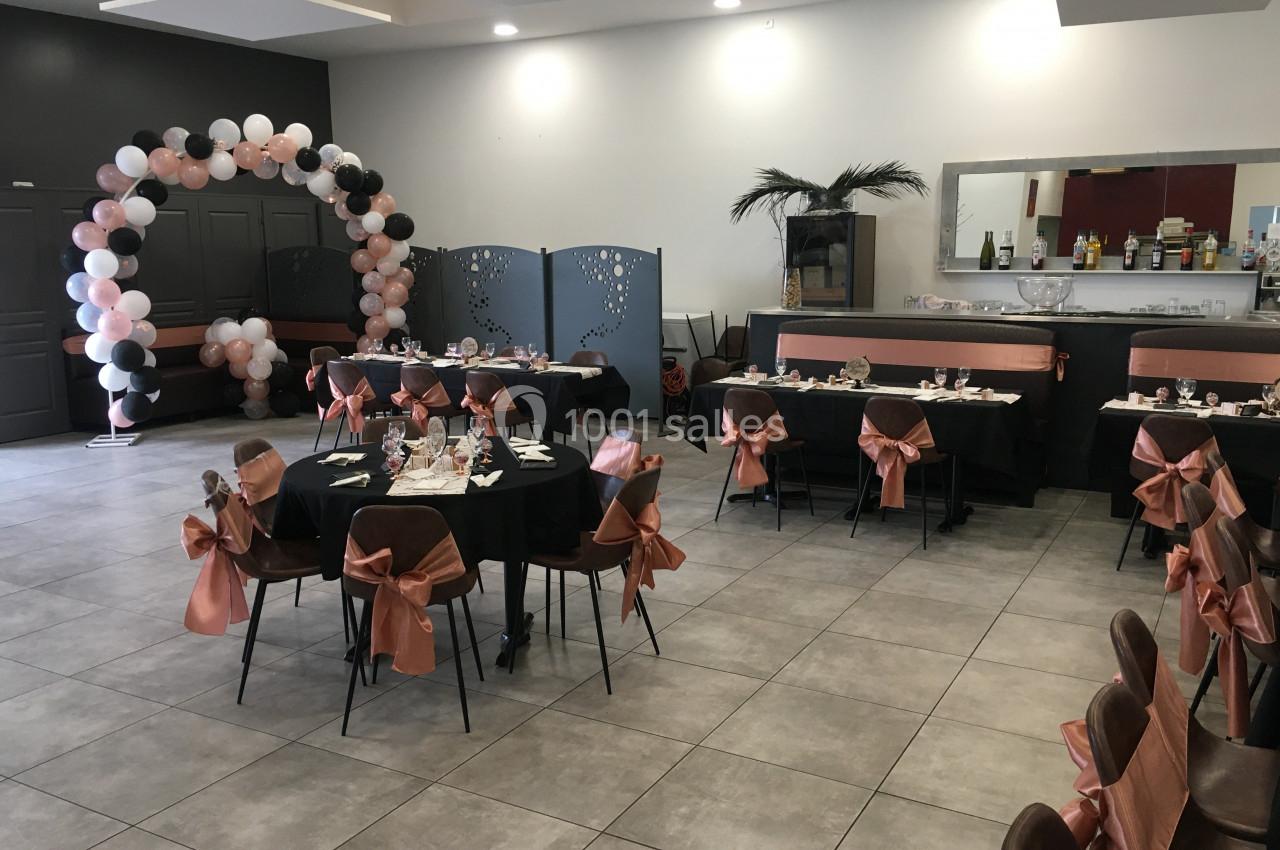 Salle décorée pour un événement, avec tables dressées, chaises ornées de nœuds roses et arche de ballons noir et rose.
