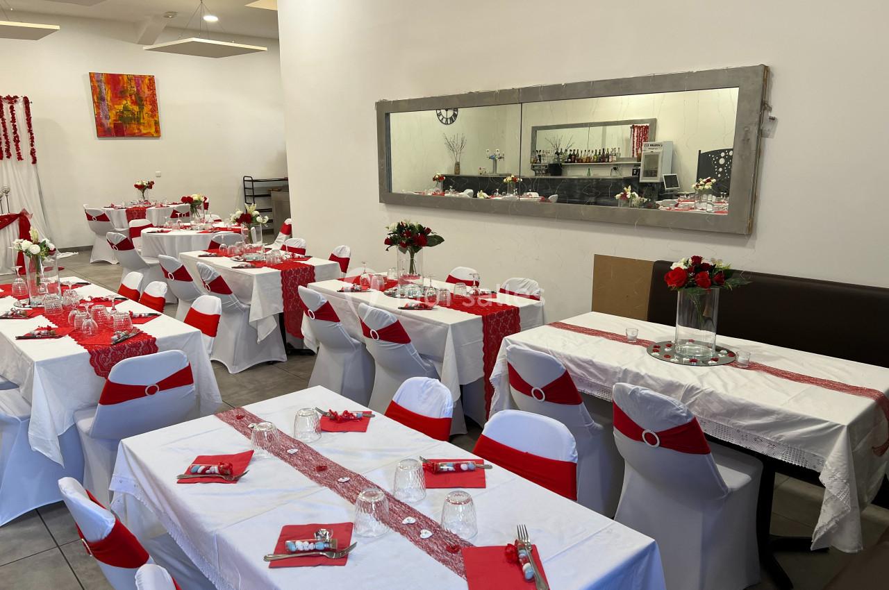 Salle de réception décorée en rouge et blanc avec des tables dressées et des chaises ornées de nœuds rouges.