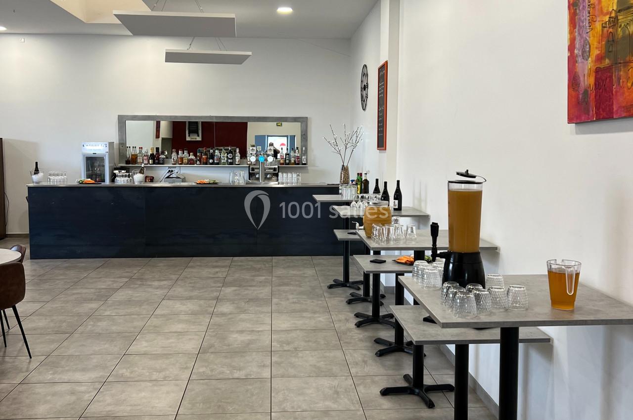 Salle avec un bar en fond, tables alignées avec verres, carafes de jus et bouteilles disposées sur la droite.