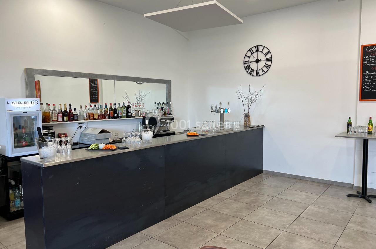 Comptoir de bar avec bouteilles alignées, verres, machine à café et décor sobre dans une pièce lumineuse.