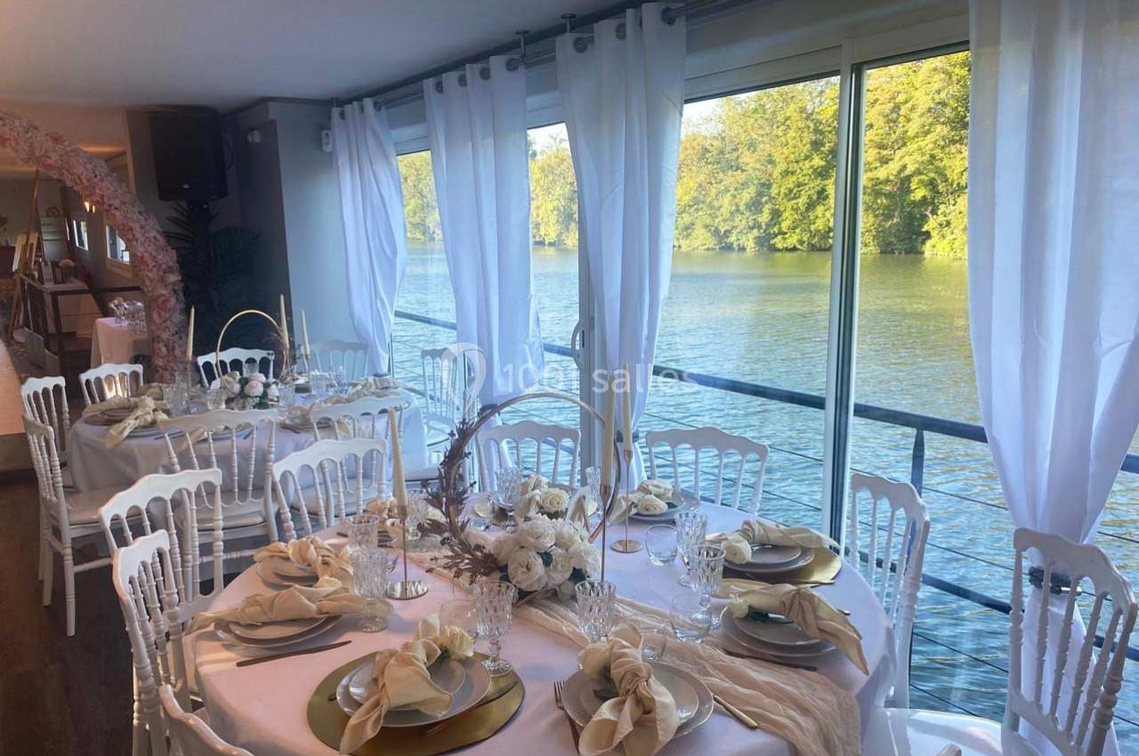 Salle de réception élégante avec tables décorées de fleurs blanches et vue sur un cours d'eau bordé d'arbres.