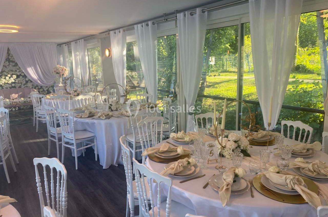 Salle de réception lumineuse avec tables décorées de nappes blanches, vaisselle élégante et chaises blanches, vue sur jardin.