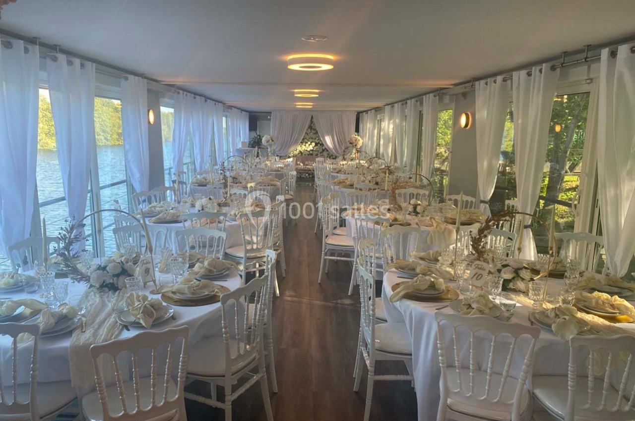 Salle de réception élégamment décorée avec des tables rondes, nappes blanches et chaises disposées près de grandes baies…
