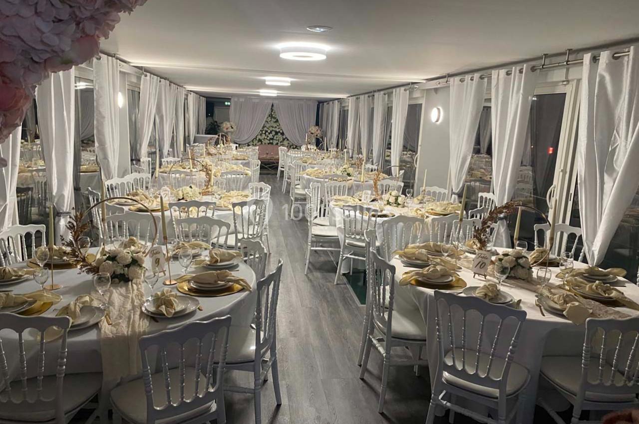 Salle de réception décorée avec des tables rondes dressées, nappes blanches, vaisselle élégante et rideaux translucides.