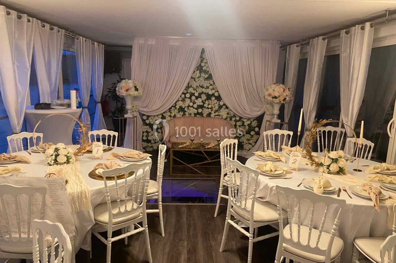 Salle décorée pour un événement avec tables dressées, chaises blanches et un fond floral blanc derrière un canapé rose.