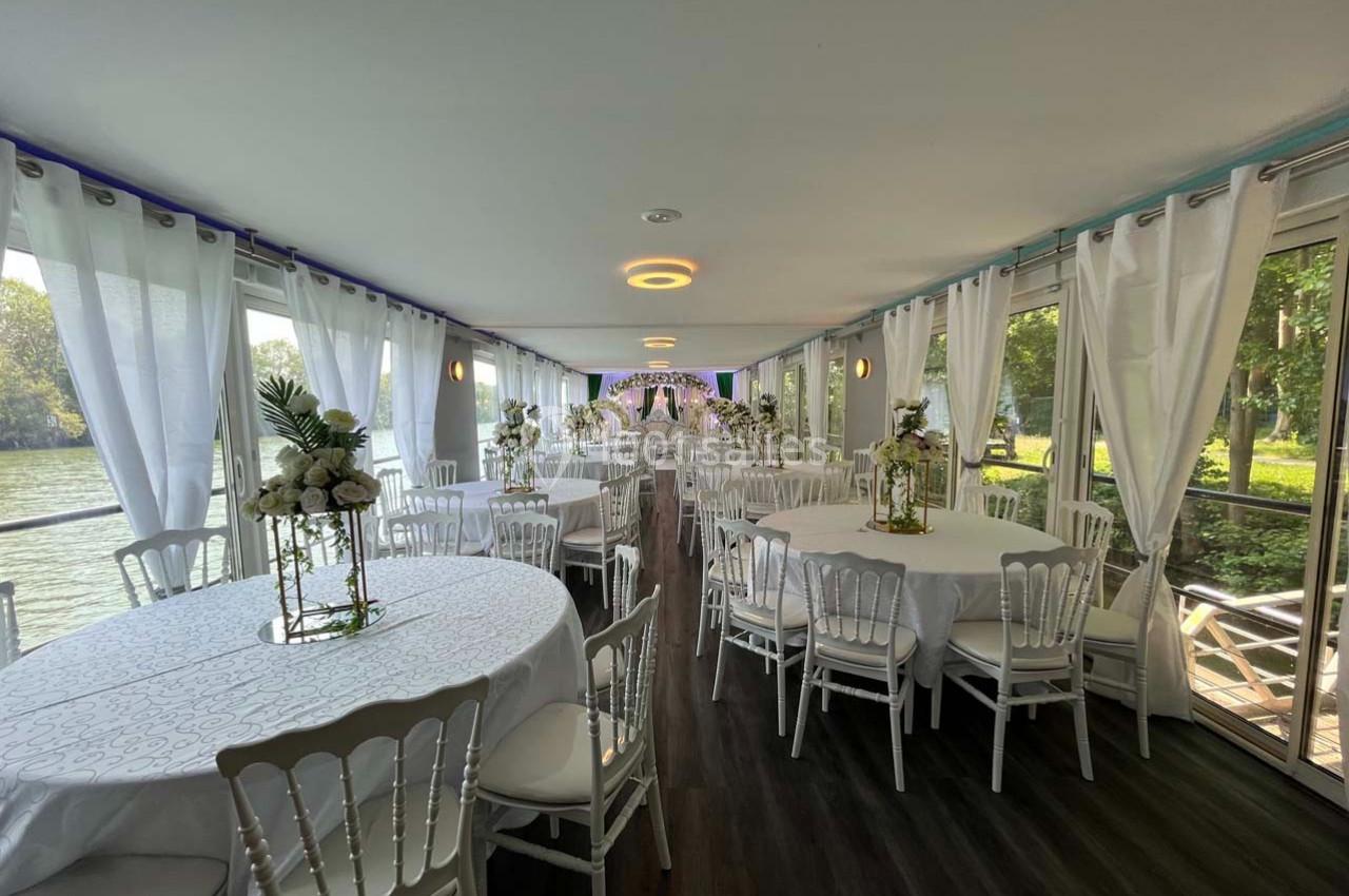 Salle lumineuse avec grandes baies vitrées, tables rondes décorées de fleurs blanches et chaises blanches élégantes.