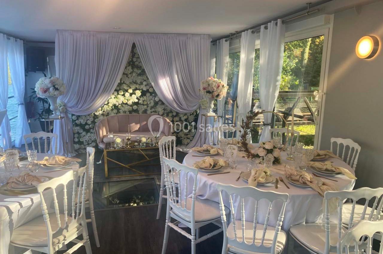 Salle décorée pour un événement avec tables dressées, chaises blanches, rideaux blancs et mur floral en arrière-plan.