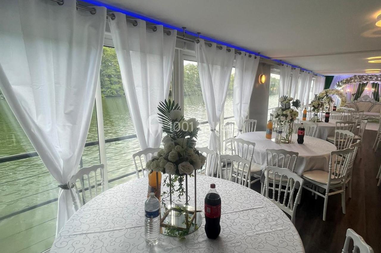 Salle de réception décorée avec des tables rondes, nappes blanches, bouquets de fleurs et vue sur un cours d'eau par de…