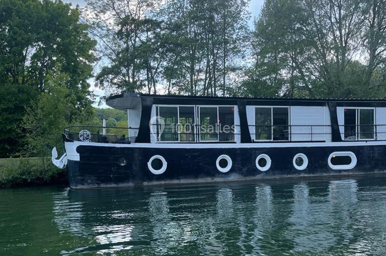 Bateau noir et blanc amarré sur une rivière, entouré d'arbres et reflété dans l'eau calme.