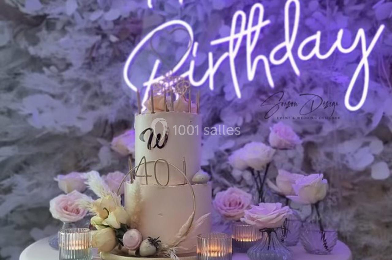 Gâteau d'anniversaire décoré de fleurs et bougies, devant un fond floral avec une enseigne lumineuse ’Happy Birthday’.