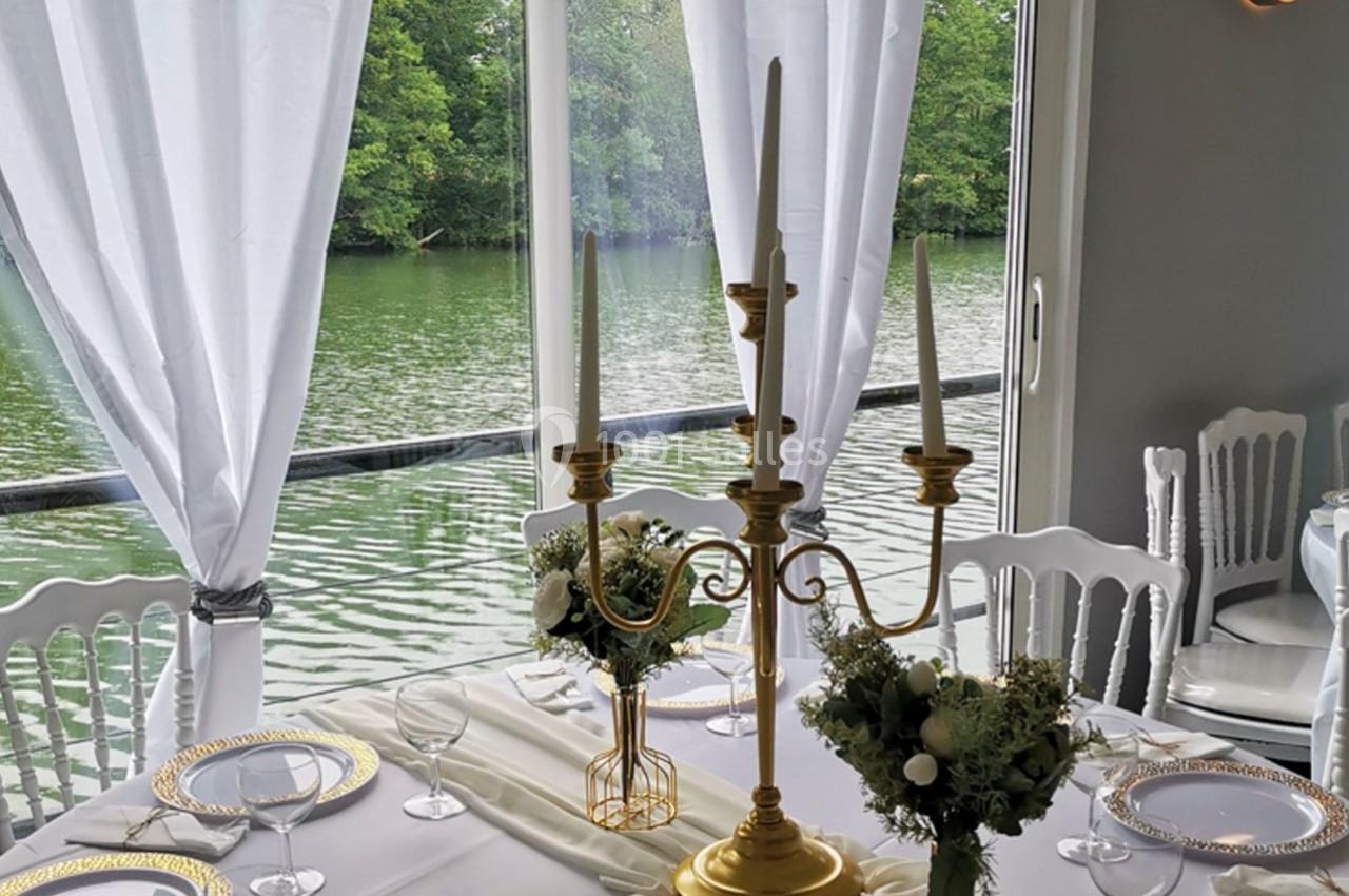 Table élégamment dressée avec chandelier doré, fleurs et vue sur un lac à travers de grandes baies vitrées.