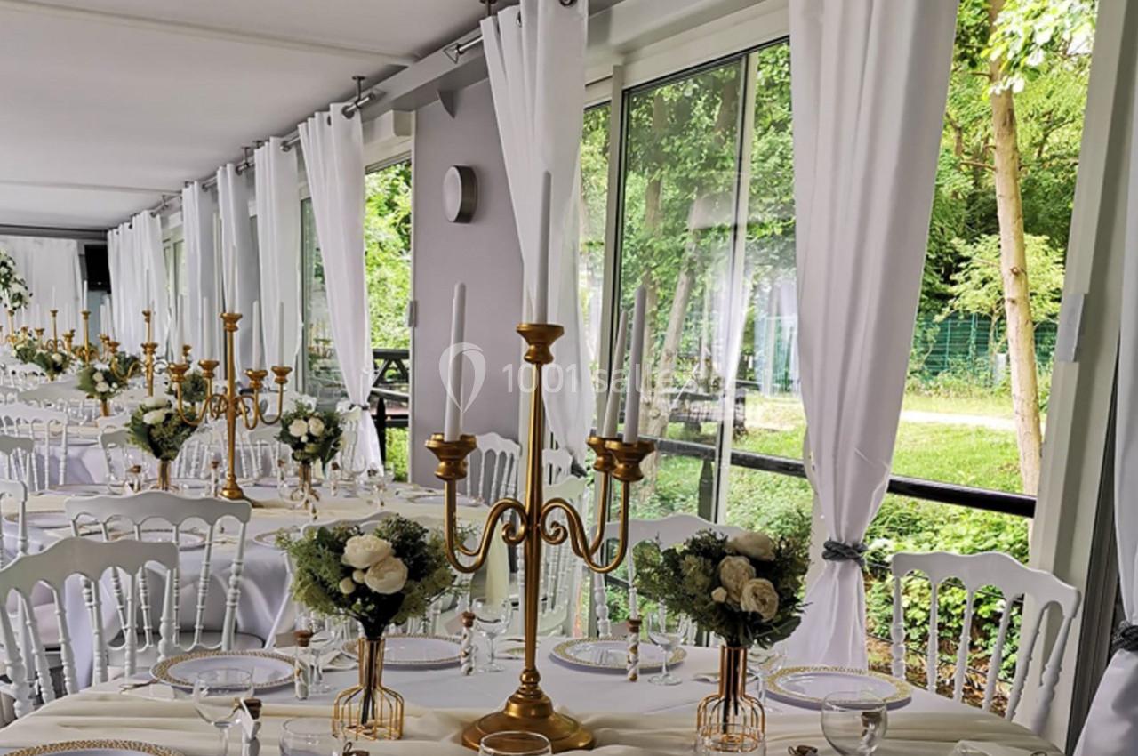 Salle de réception lumineuse avec tables décorées de chandeliers dorés, fleurs blanches et nappes blanches, vue sur jardin.