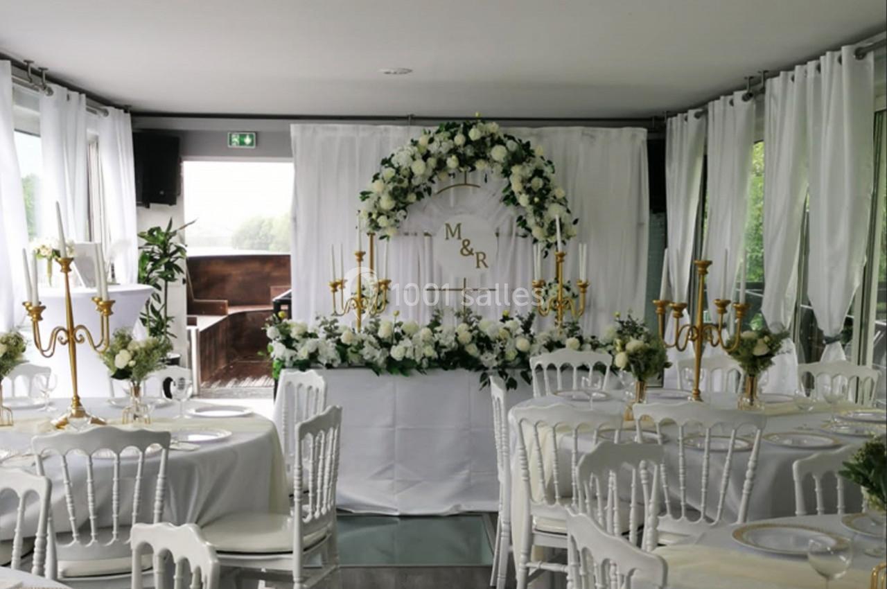 Salle décorée pour un mariage avec des tables rondes, des chaises blanches, des chandeliers dorés et une arche florale.