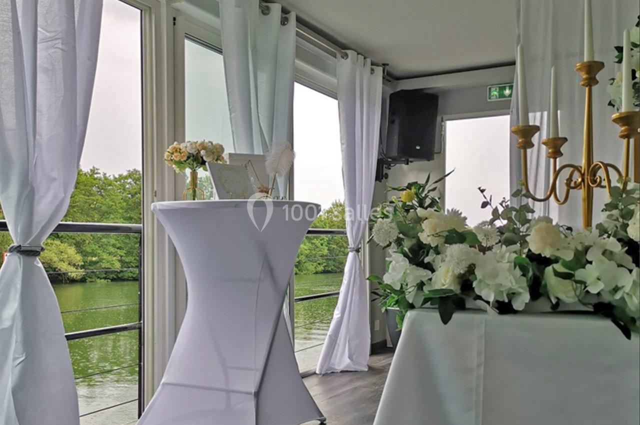 Salle lumineuse avec vue sur un plan d'eau, décorée de fleurs blanches, bougies et une table haute drapée de blanc.