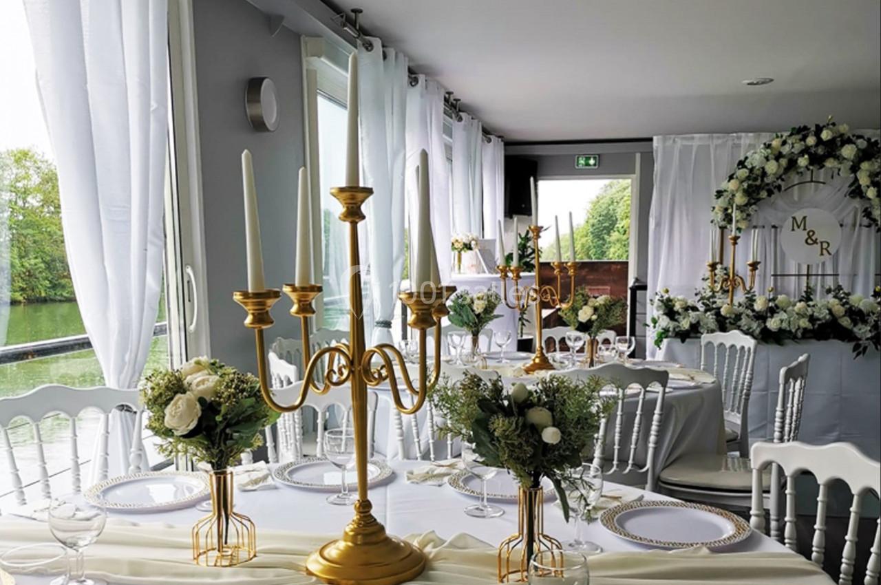Salle de réception décorée avec des chandeliers dorés, des fleurs blanches et des chaises blanches près de grandes fenêtres.