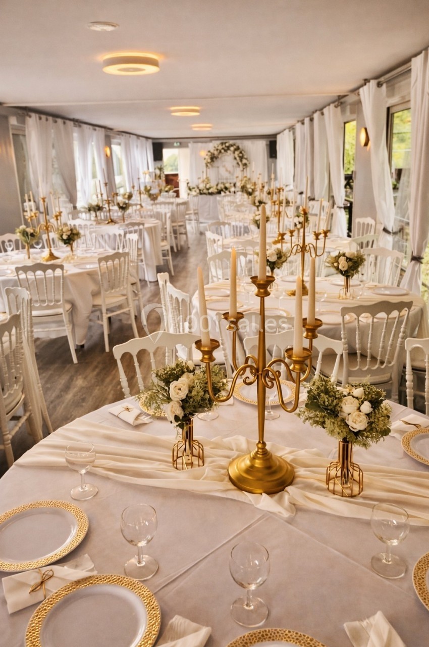 Salle de réception décorée avec des tables rondes, nappes blanches, chandeliers dorés et compositions florales.