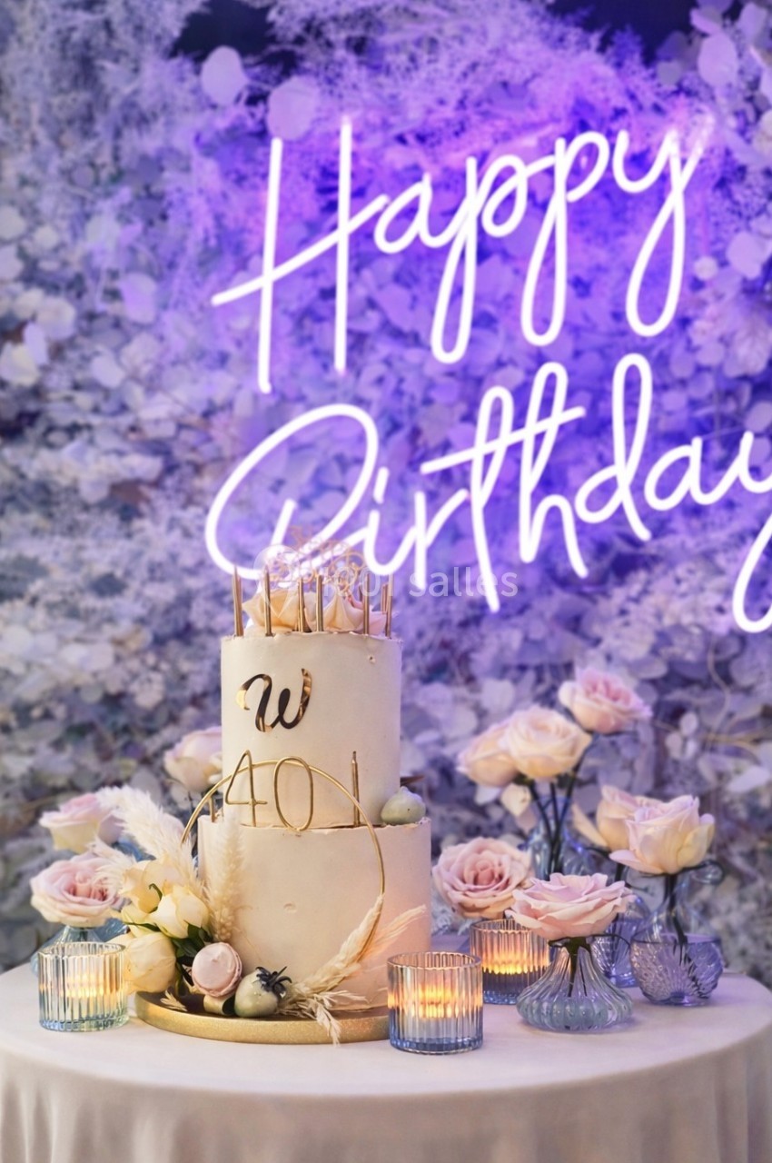 Gâteau d'anniversaire décoré de fleurs et bougies, entouré de roses, avec un néon ’Happy Birthday’ en arrière-plan.
