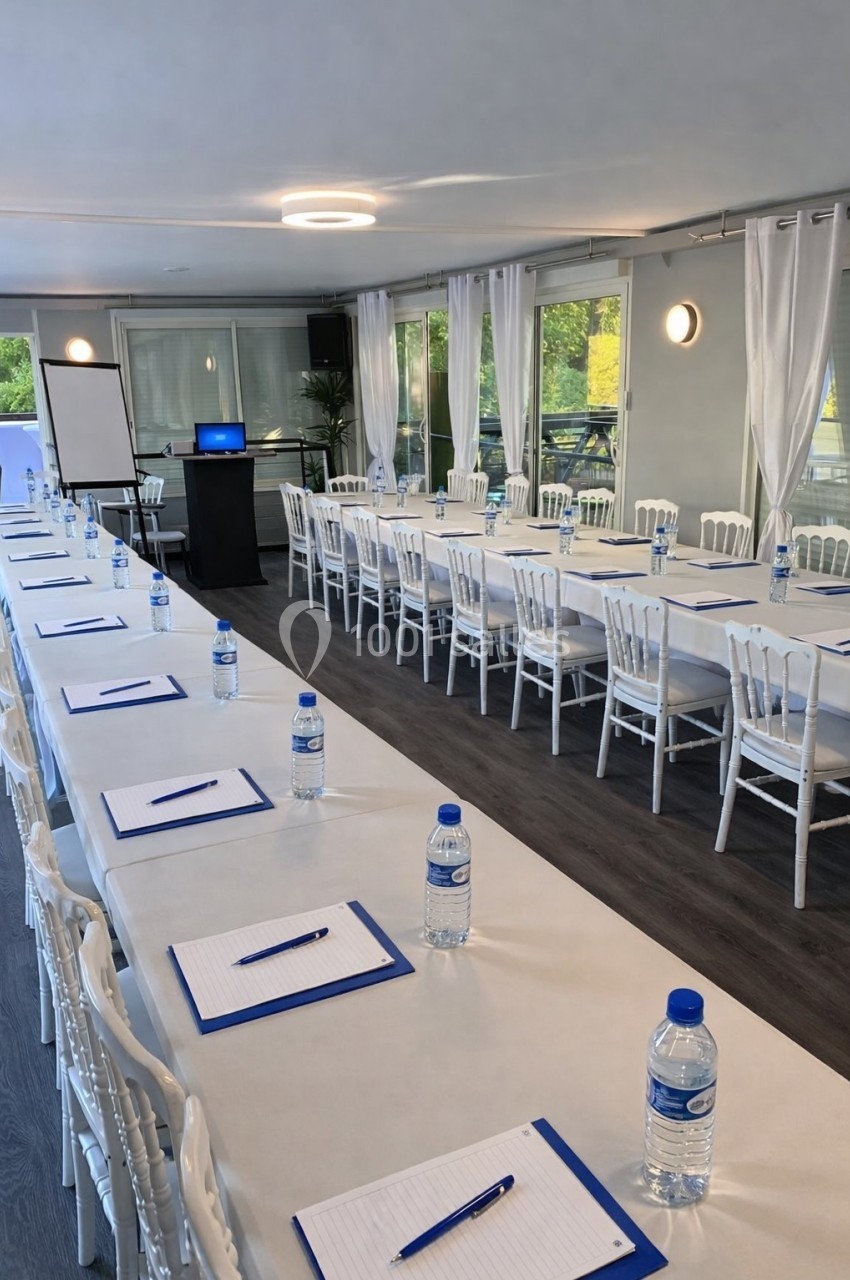 Salle de réunion lumineuse avec tables alignées, chaises blanches, blocs-notes, bouteilles d'eau et écran de présentation.