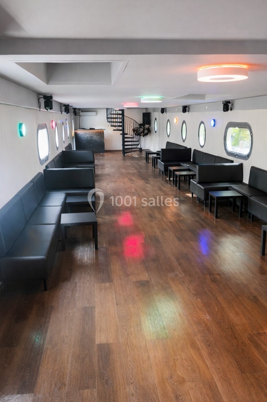 Salle intérieure lumineuse avec parquet en bois, banquettes noires, tables basses et hublots ronds sur les murs.