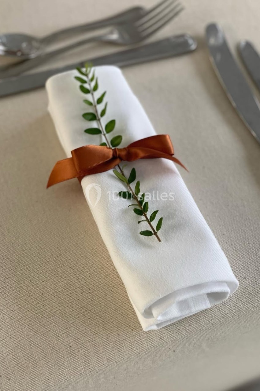 Serviette blanche roulée, décorée d'une branche verte et d'un ruban orange, posée sur une table dressée.