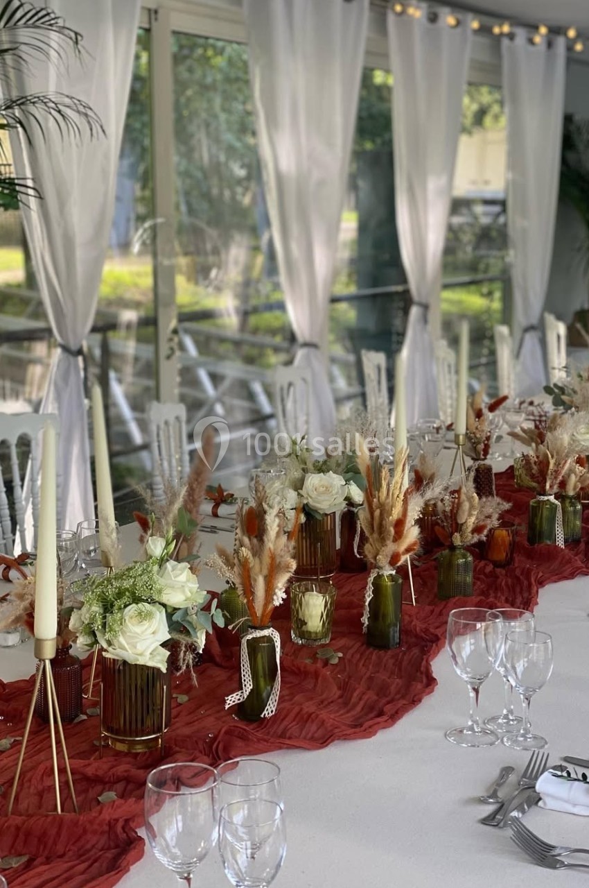 Table décorée pour un événement, avec nappes blanches, chemin de table rouge, bougies, fleurs et herbes séchées.
