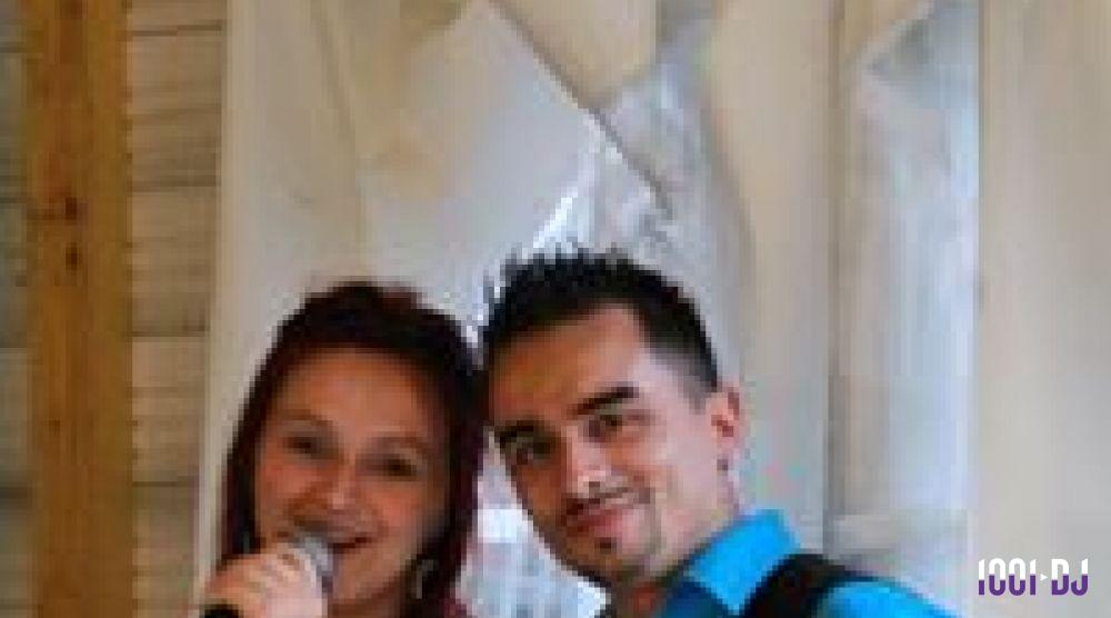 Un homme et une femme souriants, la femme tenant un micro, devant un fond clair avec des rideaux.