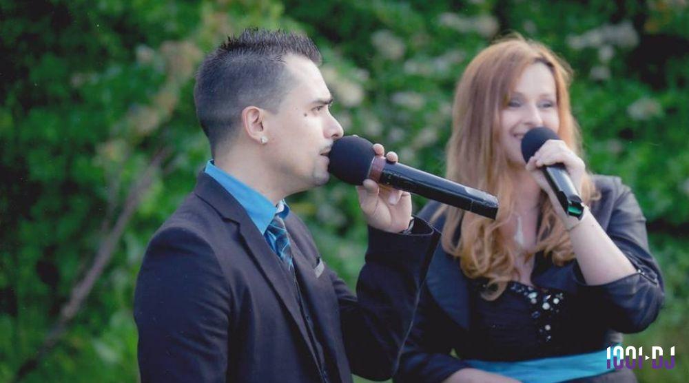 Deux personnes chantant dans des micros en extérieur, devant une végétation verdoyante.