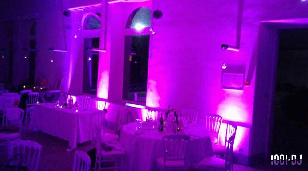 Salle de réception éclairée par des lumières violettes, avec tables dressées et chaises blanches.