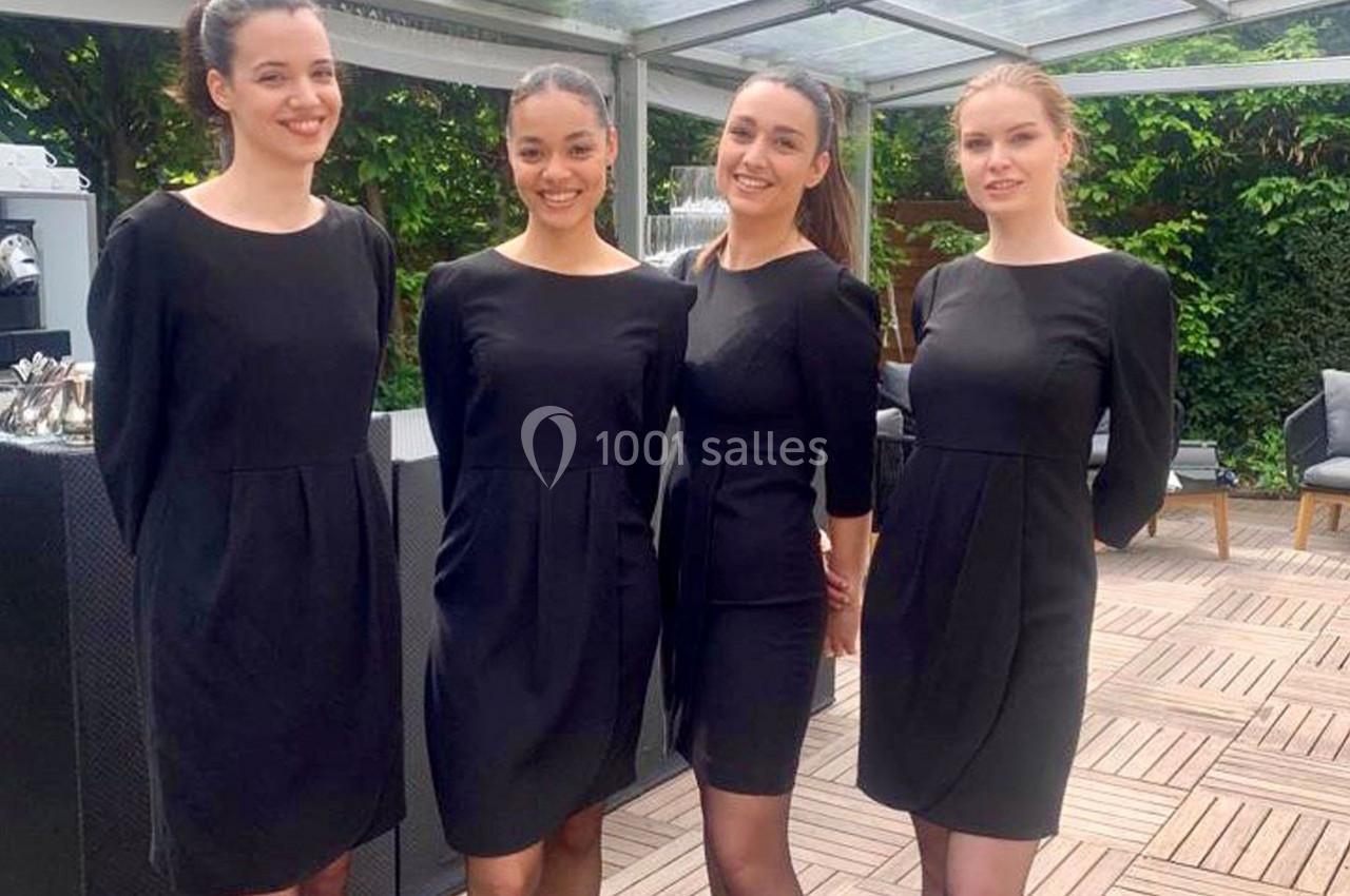 Quatre femmes en robe noire sourient debout sur une terrasse en bois avec un fond de verdure.