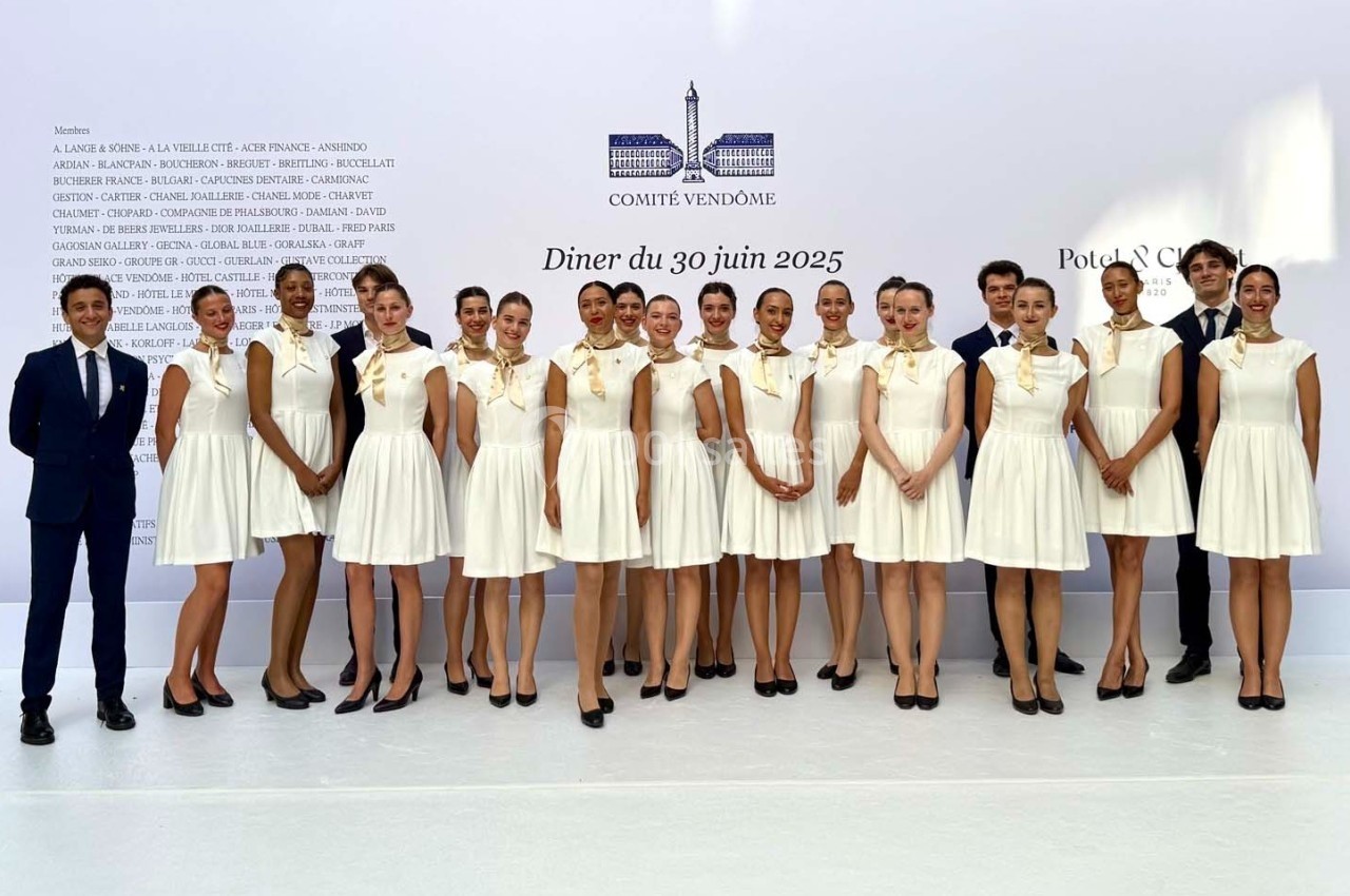 Un groupe de personnes posant en tenue élégante devant un mur blanc avec des inscriptions et un logo.