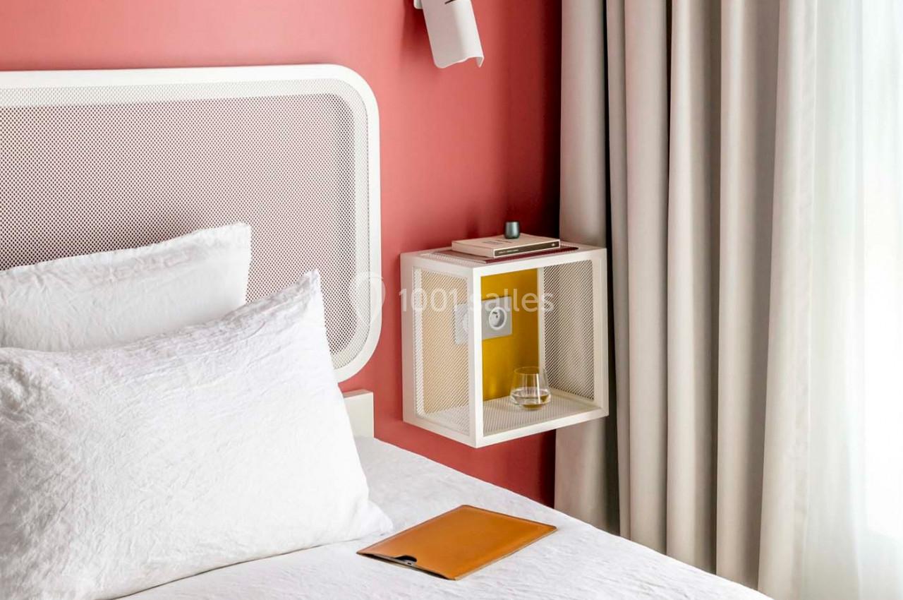 Lit avec tête de lit blanche, table de chevet murale contenant un verre et des livres, rideaux et mur rouge en arrière-plan.