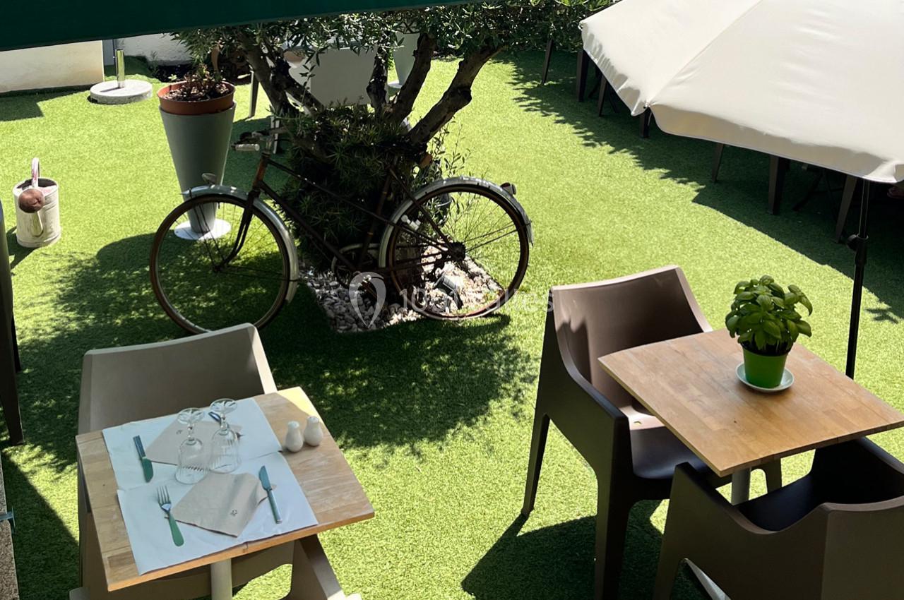 Terrasse avec tables en bois, chaises en plastique, vélo décoratif près d'un olivier et parasols sur pelouse synthétique.