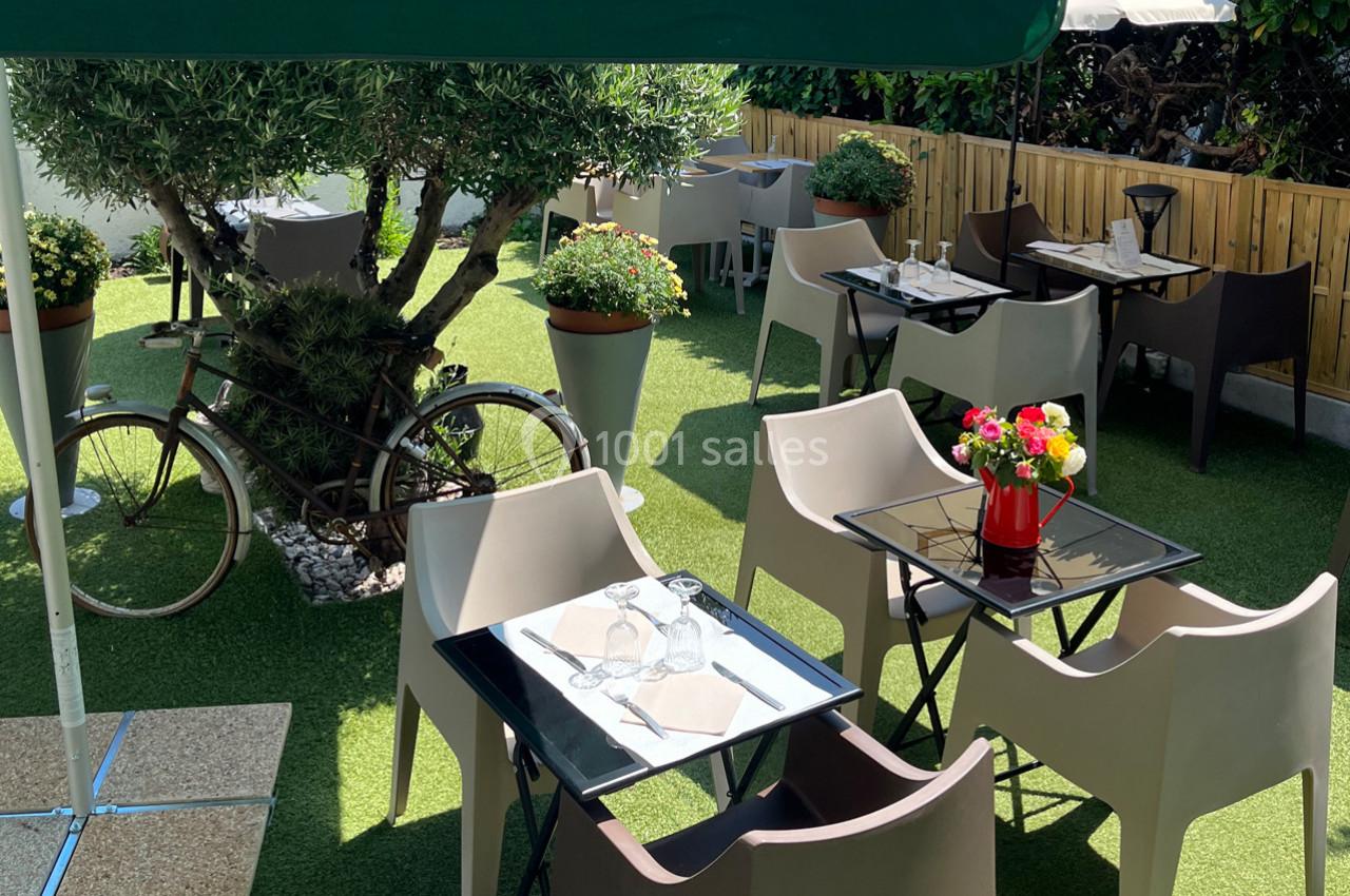 Terrasse extérieure avec tables, chaises, plantes en pot, vélo décoratif et sol en gazon synthétique.