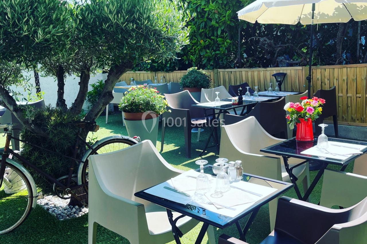 Terrasse extérieure avec tables dressées, chaises en plastique, parasol, plantes et vélo appuyé contre un arbre.