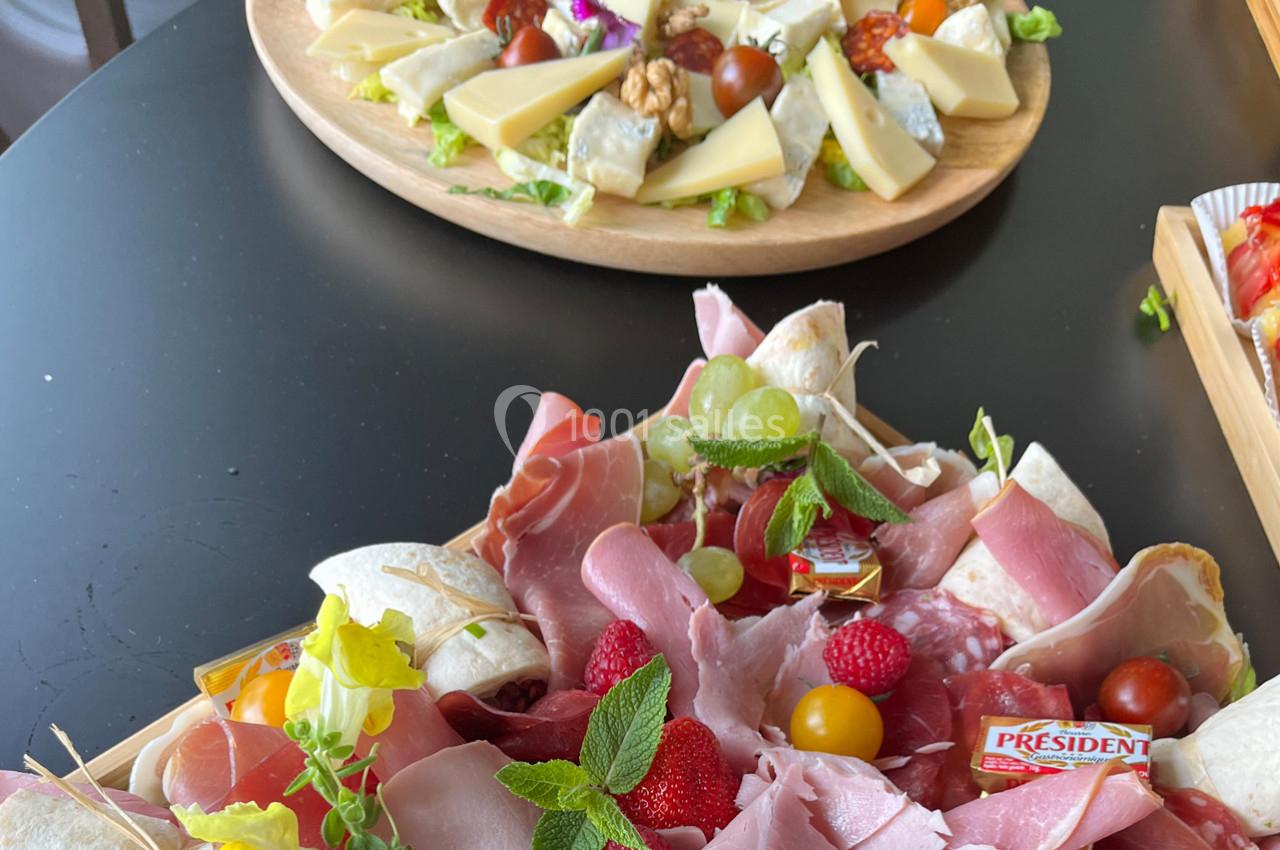 Plateau de charcuterie avec fruits et herbes fraîches au premier plan, plateau de fromages variés en arrière-plan.