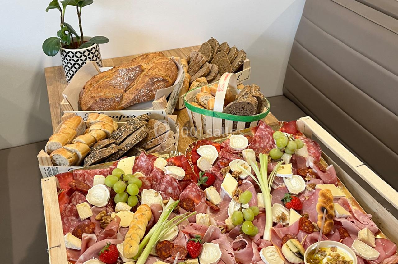 Plateau de charcuterie, fromages, fruits et pains variés disposés sur une table avec une plante en arrière-plan.